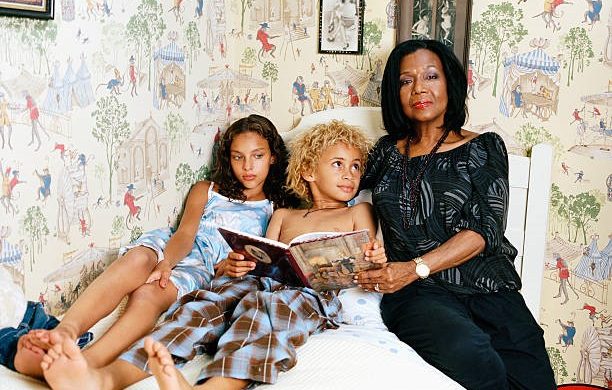 An older woman sitting on a bed with two children, reading a book together.