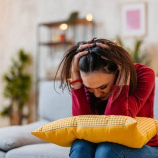 A stressed woman sitting on a couch with a pillow in her lap, holding her head.