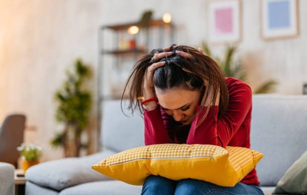 A stressed woman sitting on a couch with a pillow in her lap, holding her head.