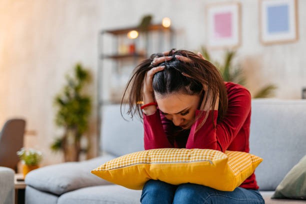 A stressed woman sitting on a couch with a pillow in her lap, holding her head.