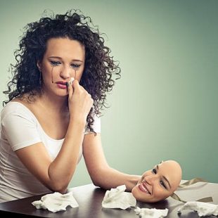 A lady wiping her tears with one hand and holding a smiling mask from another while sitting in front of a table.