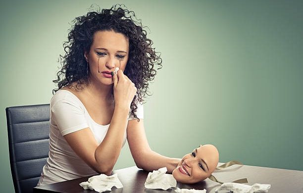 A lady wiping her tears with one hand and holding a smiling mask from another while sitting in front of a table.