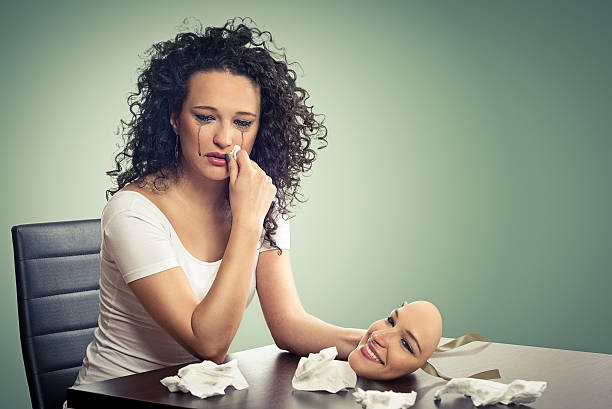 A lady wiping her tears with one hand and holding a smiling mask from another while sitting in front of a table.