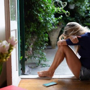 A lady sitting in an open door’s frame, hugging her knees with a phone lying beside her on the floor.