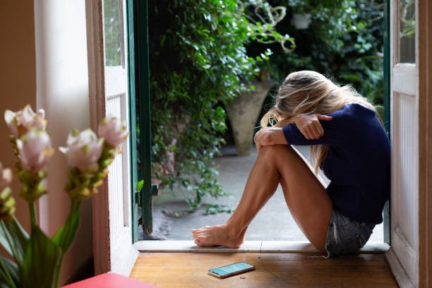 A lady sitting in an open door’s frame, hugging her knees with a phone lying beside her on the floor.